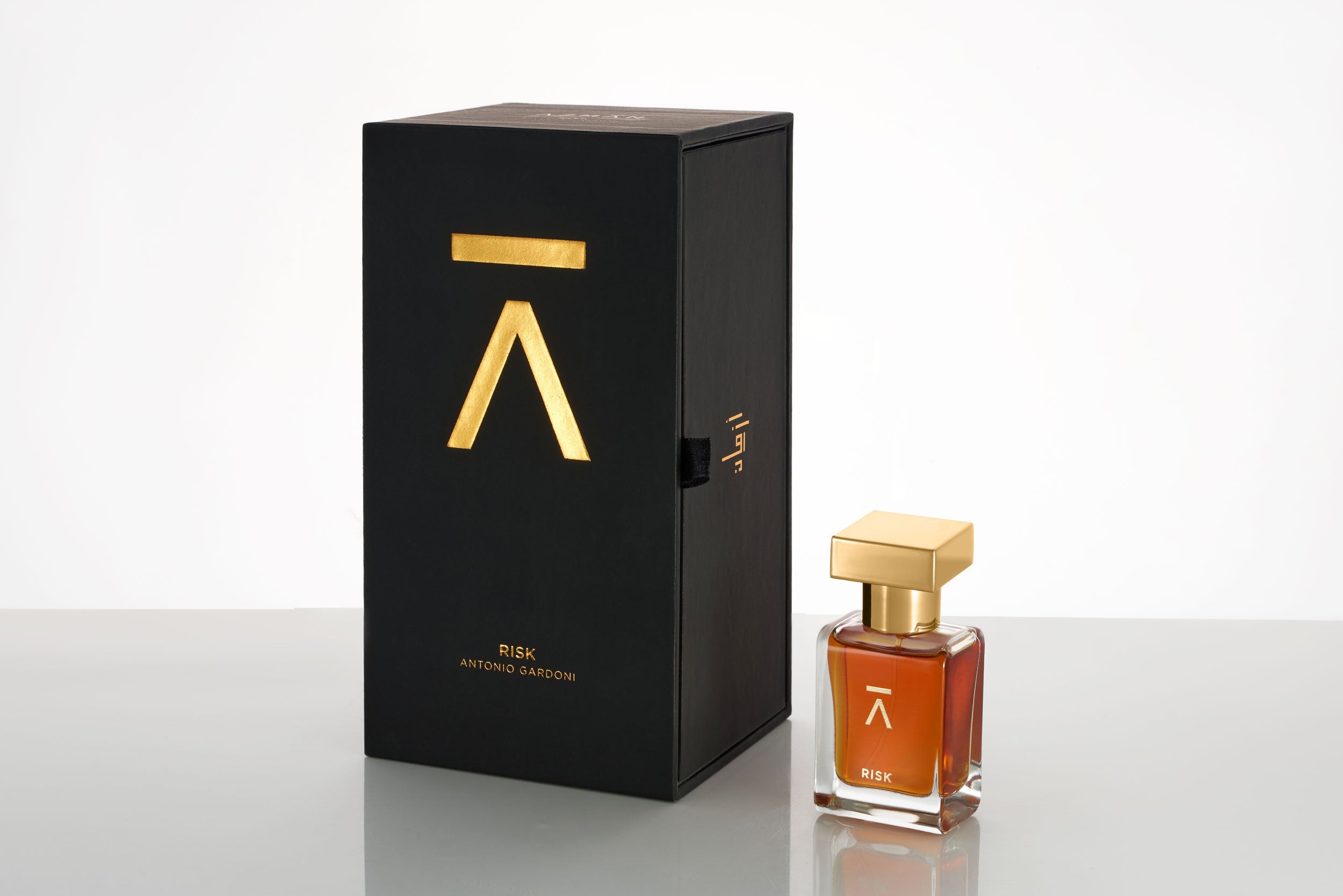 Azman Risk Perfume collaboration with Italian perfumer Antonio Gardoni. Majnoon bottle features a long neck, hand-polished bottle with a heavy gold metal cap. The packaging includes a side-opening heavy cardboard box adorned with a gold foil Azman logo. The studio shot of the Risk perfume bottle is set against a white color background. The whisky gold color of the Risk perfume adds a touch of luxury and sophistication